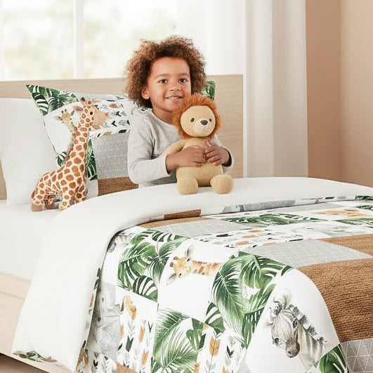 Enfant souriant jouant avec une peluche lion dans un lit simple couvert par un ensemble de literie patchwork safari jungle. La couverture présente des motifs d'animaux africains (lionceau, zèbre, girafe) et de feuilles tropicales. Scène lifestyle chambre enfant.
