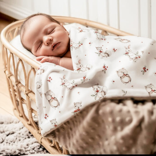 Couverture bébé minky réversible "Canneberges et Petites Pattes" avec motif animaux de la forêt (lapin, hérisson), utilisée pour envelopper un nouveau-né endormi dans un berceau en rotin. Le revers taupe texturé en minky est visible, soulignant la double douceur. Photo lifestyle lumineuse et chaleureuse